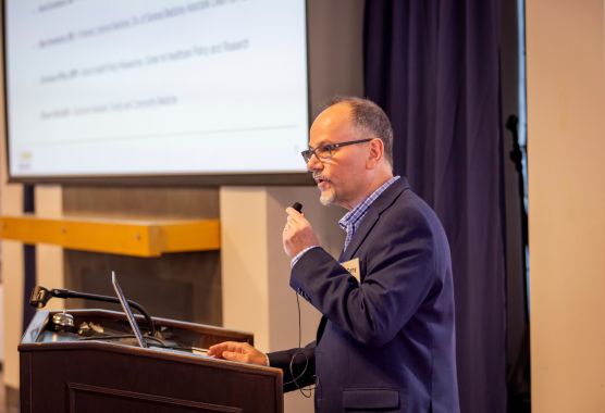 A person wearing glasses and a suit jacket speaks into a microphone at a podium with a laptop and a projector screen behind him.