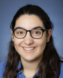 A person with long, dark hair smiles in-front of a blue background, wearing a blue shirt and glasses.