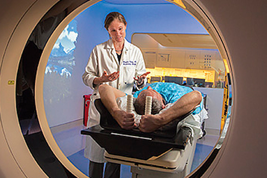 A physician in a white coat speaks with a patient being positioned in a medical imaging scanner.