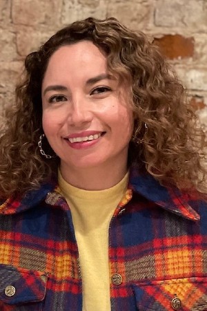Headshot of Mayra smiling in of red brick wall