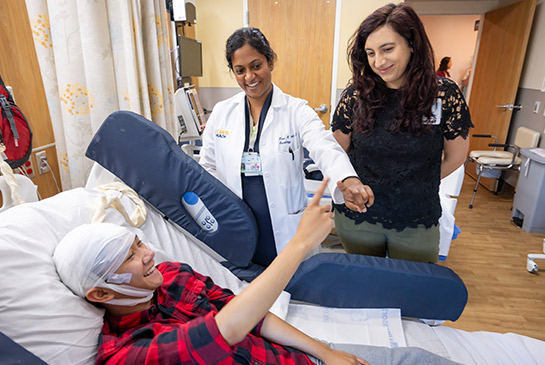 Smiling patient in hospital bed next to neurologists Kiran Kanth and Melissa Asmar.