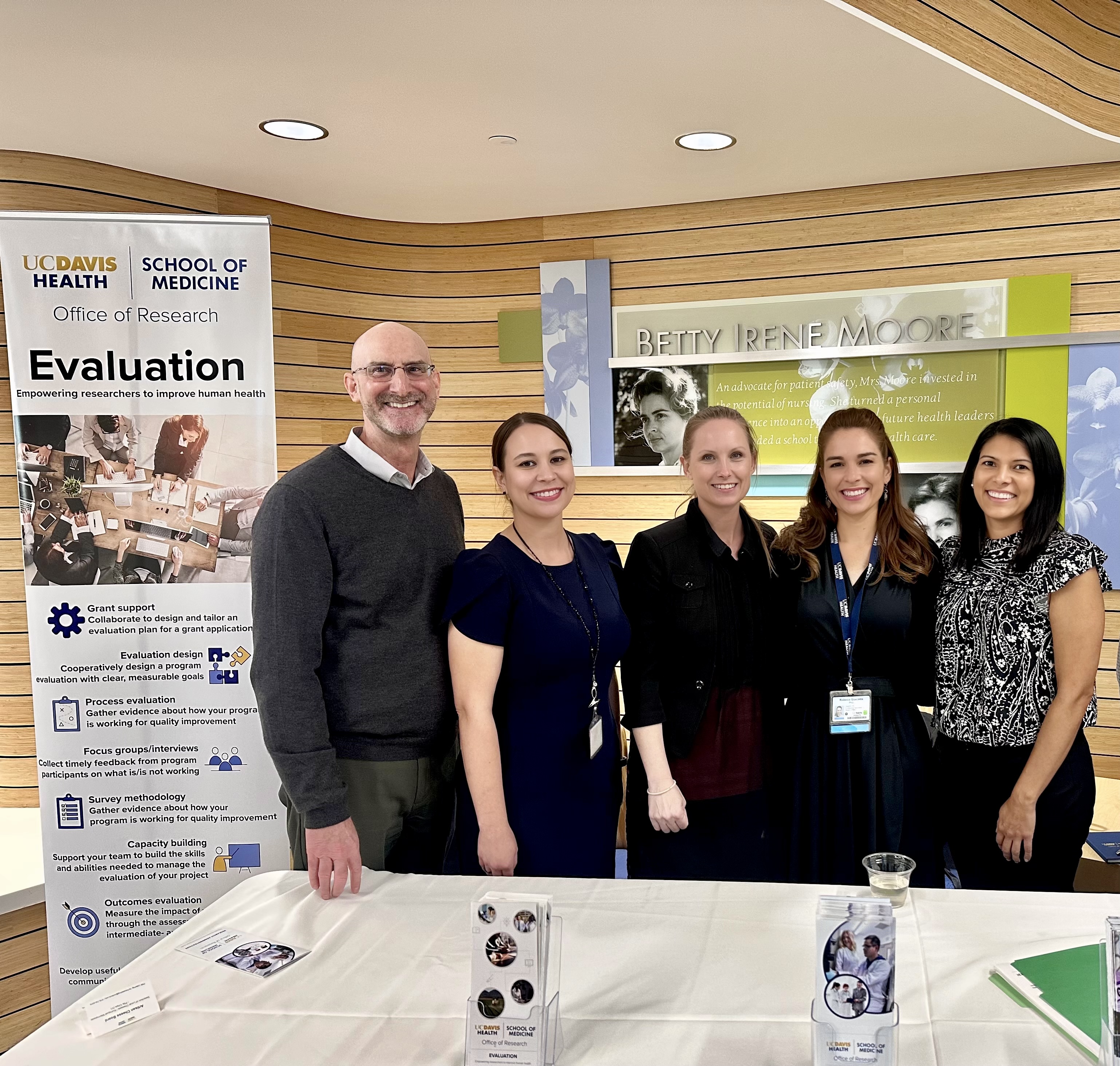 five professionals at a conference standing behind a table, next to a banner that reads Evaluation.