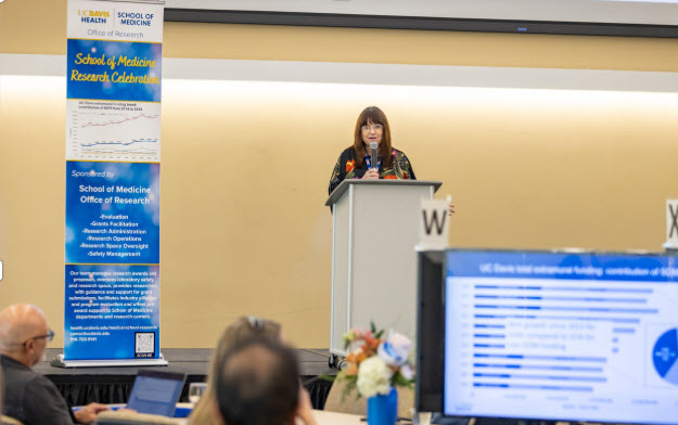 a woman stands behind a podium next to a banner that says School of Medicine Research Celebration; in the foreground is a computer monitor display