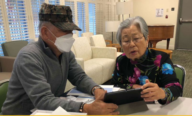 Two older people in a common room, one wearing a mask, look at a computer tablet together