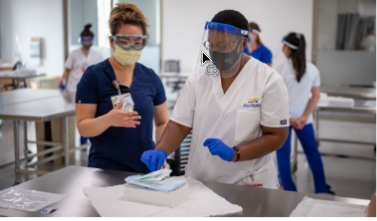 Two health professionals, one in blue scrubs and one in white scrubs, both wearing personal protective gear, work carefully with medical equipment
