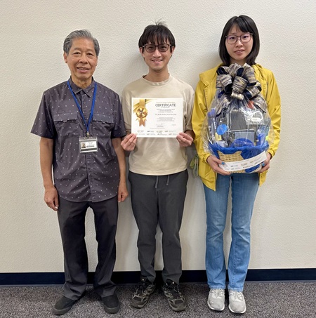 Three individuals pose in a hallway, the middle individual holding a certificate
