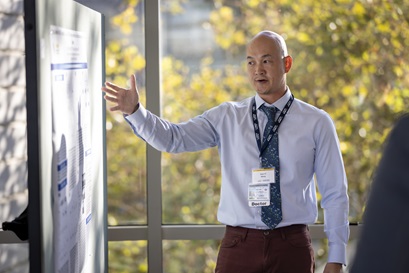 a professionally dressed man gives a poster presentation in front of a large window
