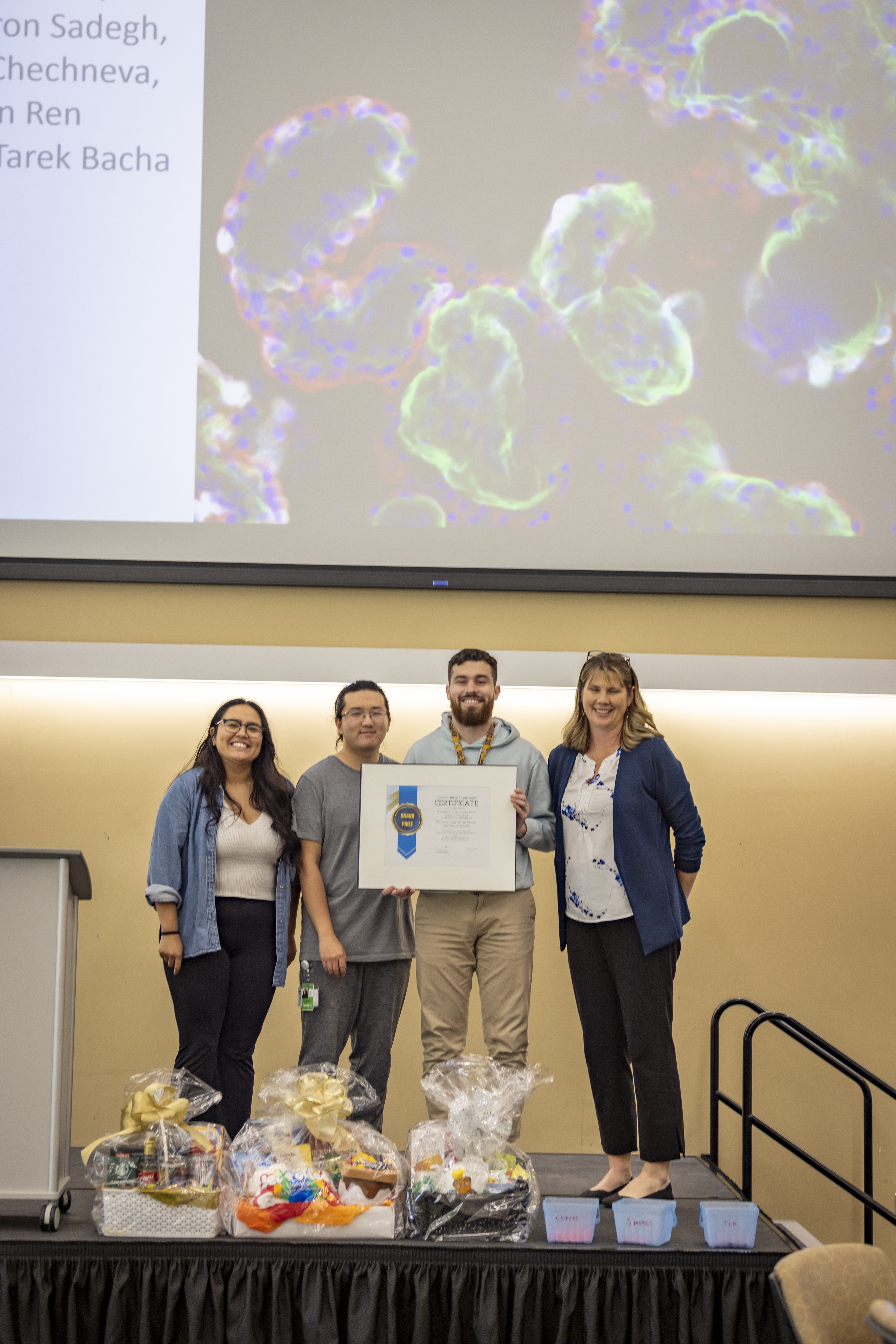 four people on a stage, with a large projection behind them displaying a microscopy image; in the foreground are gift baskets; the third individual to the left holds a large framed certificate
