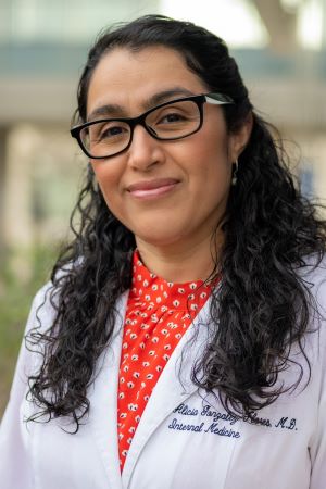 Portrait of Alicia Gonzales-Flores wearing a white lab coat over a red patterned blouse, outdoors with greenery blurred in the background.