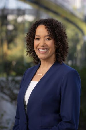 Portrait of Brandee Waite wearing a blue blazer and white top, standing outdoors with campus structures and greenery softly blurred in the background.