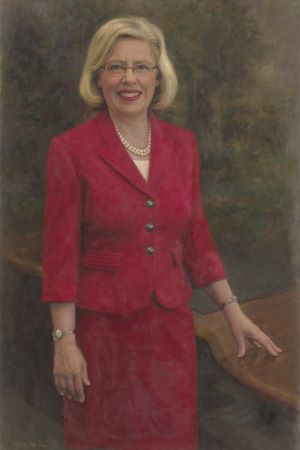 Formal painted portrait of Claire Pomeroy wearing a red suit and pearl necklace, standing beside a table against a softly blended, neutral background.