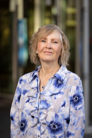 Portrait of Colleen Sweeney wearing a blue floral blouse standing outdoors in front of a glass building with a softly blurred background.
