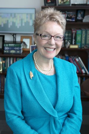 Portrait of Diana Farmer wearing a blue jacket and pearl necklace, seated in an office setting with bookshelves and framed photos visible in the backg