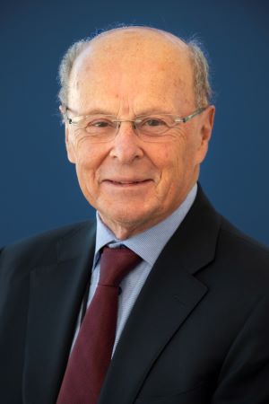 Studio portrait of Fred Meyers wearing a dark suit jacket, light blue shirt, and burgundy tie, photographed against a solid blue background.