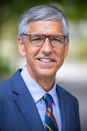 Portrait of Mark Henderson wearing a blue suit jacket, light blue shirt, and multicolored striped tie, photographed outdoors.