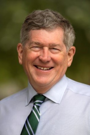 Portrait of Mark Servis wearing a light blue dress shirt and green striped tie, photographed outdoors with greenery softly blurred in the background.
