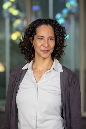 Portrait of Micaela Godzich wearing a light button-down shirt and dark cardigan, standing indoors with a softly blurred background.