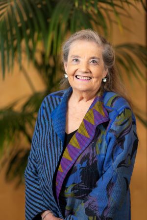 Portrait of Randi Hagerman wearing a patterned blue and purple shawl over a dark top, standing indoors with palm leaves and a warm-toned background.