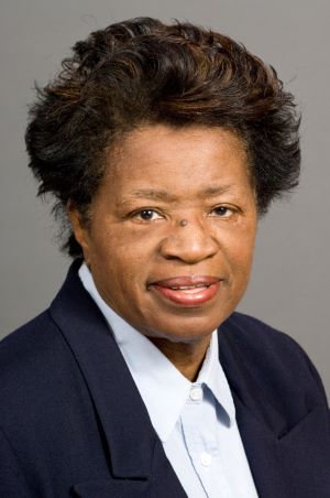 Studio portrait of Sarah Gray wearing a dark blazer over a light collared shirt, photographed against a plain neutral background.