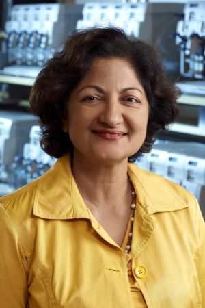 Portrait of Satya Dandekar wearing a yellow jacket, standing in a laboratory setting with scientific equipment visible in the background.