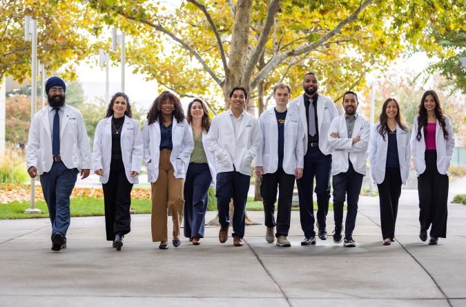 medical students in white lab coats walk together along a tree‑lined campus pathway with autumn foliage, smiling and talking