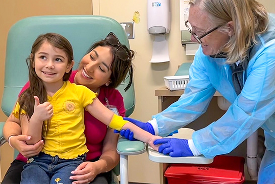 mom and child sitting in a chair with a phlebotomist giving a blood draw
