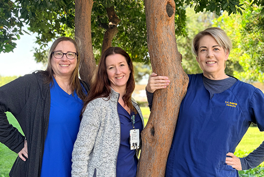 Three MIND Institute nurses standing together outside near a tree, smiling and wearing blue scrubs and ID badges