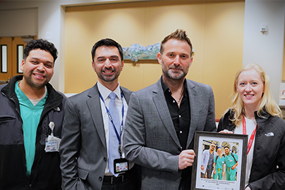 Three men and a woman pose while two of them hold a picture that shows two residents in green scrubs and a doctor in a white coat