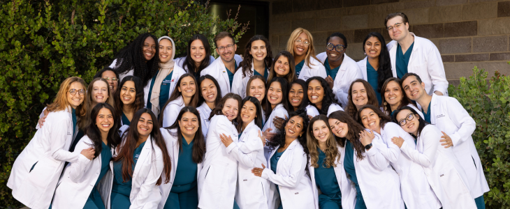 Group photo of OBGYN residents smiling outdoors and wearing white medical coats and scrubs.