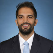 A person with dark hair and a beard in a black suit with a blue tie.