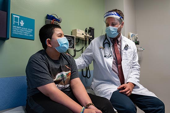 Doctor talks to pediatric patient on exam table with masks on.