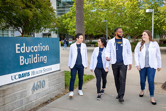 Four medical students, two males and two females, walk along the path of the UC Davis Education Builiding in white coats