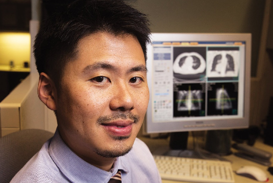 Radiation oncologist sits at a desk reviewing medical imaging scans on a large computer monitor.