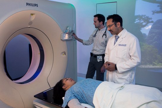 A patient lying on a table beside a large CT scanner while two medical professionals stand nearby, one adjusting the machine and the other observing.