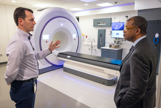 Two radiation oncology professionals standing in a modern imaging room beside an imaging and treatment machine.