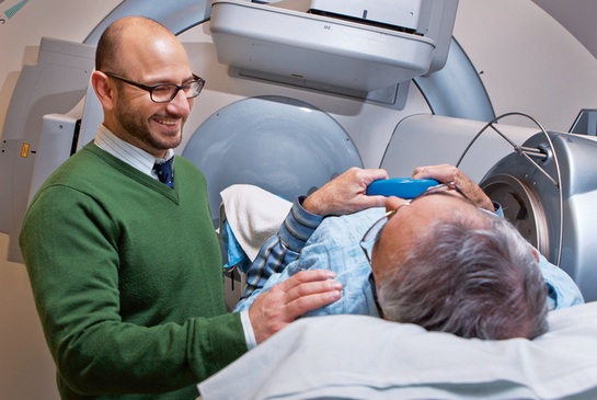 A patient lying on a medical imaging table inside a scanner while a clinician stands nearby, placing a reassuring hand on the patient’s shoulder.