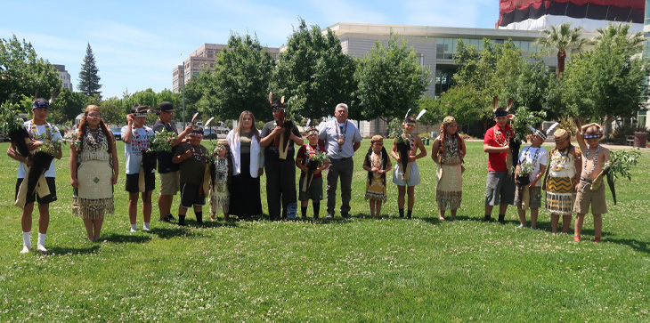 Group of tribal community members, elders and youth stand together outdoors on a grassy lawn wearing traditional regalia and holding cultural items during a demonstration.